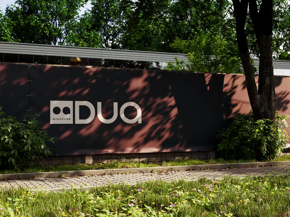 Logotipo da DUA Marketing em creme aplicado num painel escuro de rua, à sombra das árvores