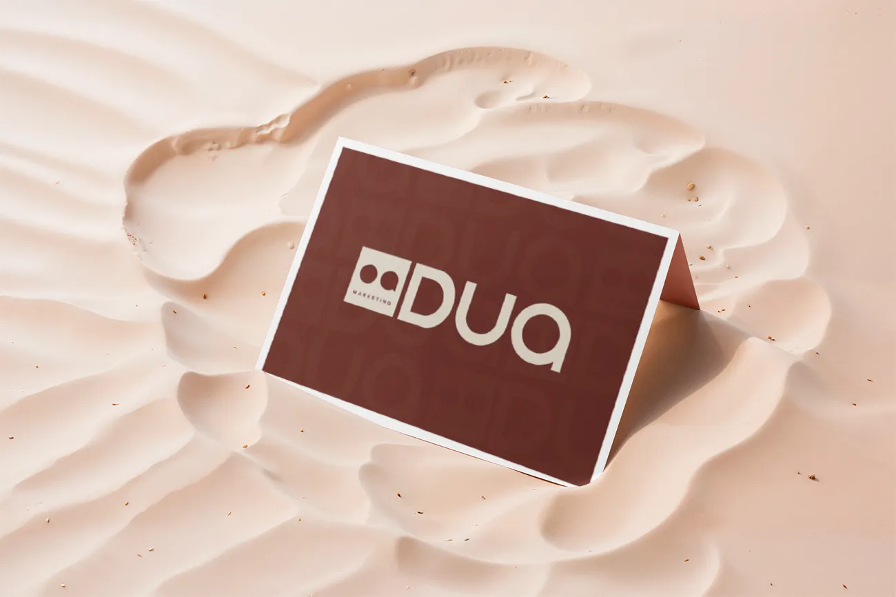 Cartão da DUA Marketing em vinho com o logotipo em creme, apoiado na areia